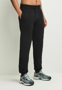 Schwarze Jogginghosen mit einem elastischen Taillenbund, Seitentaschen und schmal zulaufenden Bündchen, kombiniert mit Sportschuhen in Grau und Grün.