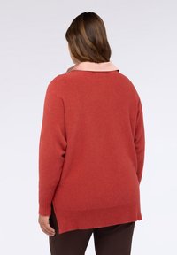 Maglione color ruggine lavorato a maglia con una texture morbida, spacchi laterali e un colletto di colore chiaro a contrasto. Indossato sopra pantaloni marrone scuro.