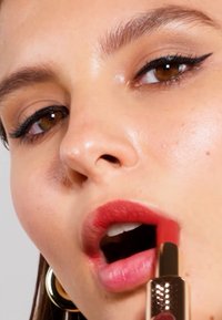 Rurka z szminką w dłoni, nakładająca głęboki matowy czerwony odcień na pełne usta. Zbliżenie podkreśla gładką teksturę i złote metaliczne opakowanie.