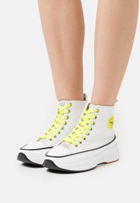 Witte high-top sneakers met chunky zolen, voorzien van neongele veters, een zilveren gestreept accent en een ronde logopatch aan de zijkant.