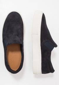 Sapatos slip-on em camurça azul-marinho com painéis laterais elásticos, sola de borracha branca e um acabamento texturizado. A palmilha apresenta uma marcação em relevo.