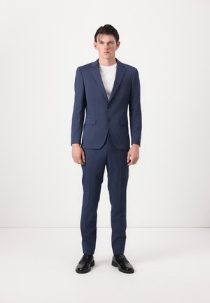 Completo su misura blu navy realizzato in tessuto strutturato, con giacca monopetto a due bottoni e pantaloni slim-fit, abbinato a una camicia bianca.