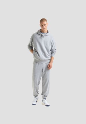 Felpa grigia con cappuccio e cordini, abbinata a pantaloni jogger coordinati. Presenta polsini a coste e sneaker casual con accenti di colore.