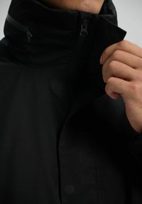 Chaqueta negra con cuello alto, cremallera completa y textura suave. Los detalles notables incluyen un bolsillo con cremallera en la parte delantera.