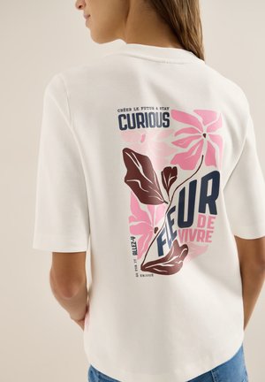 Femme portant une chemise blanche avec un motif floral rose et marron et le texte « Curious Fleur de Vivre » imprimé au dos.