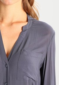 Blouse grise à col, avec boutons, dotée d'une poche poitrine et d'une texture lisse, montrée des épaules vers le haut.