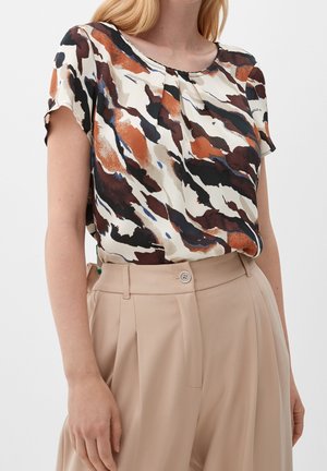 Femme portant un chemisier à manches courtes avec un motif abstrait marron, noir, blanc et orange, rentré dans un pantalon beige plissé taille haute.