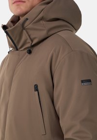 Brun vattentät jacka med hög krage och justerbar huva. Har en bröstficka med dragkedja och en logga på ärmen. Slät textur.