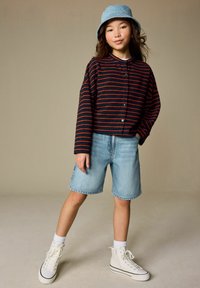 Kind draagt een navyblauw en rood gestreept overhemd met knopen, lichte denim shorts, witte hoge sneakers en een blauwe bucket hat.