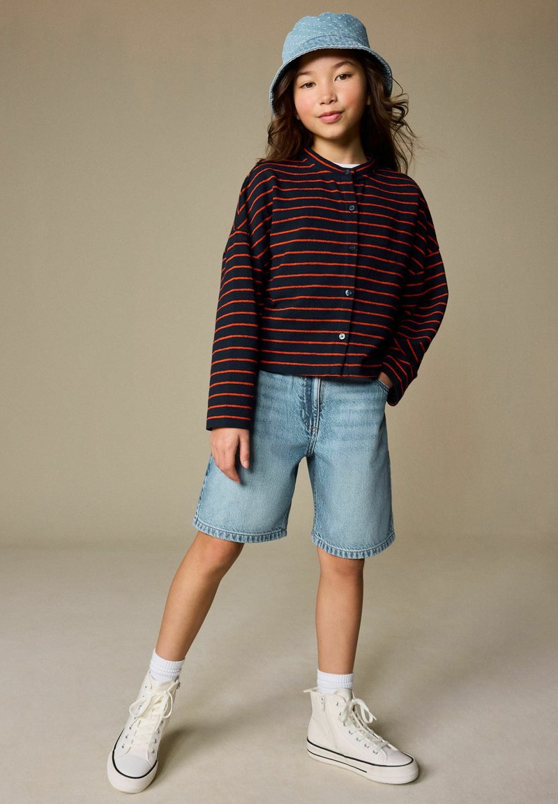 Kind draagt een navyblauw en rood gestreept overhemd met knopen, lichte denim shorts, witte hoge sneakers en een blauwe bucket hat.