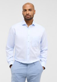 Camicia azzurro chiaro con taschino sul petto, maniche lunghe e un tessuto liscio e leggermente lucido. Caratterizzata da semplici bottoni bianchi e una vestibilità su misura.