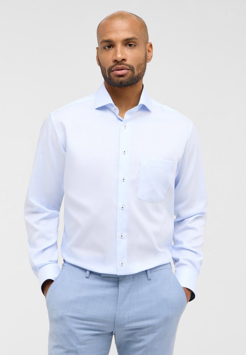 Camicia azzurro chiaro con taschino sul petto, maniche lunghe e un tessuto liscio e leggermente lucido. Caratterizzata da semplici bottoni bianchi e una vestibilità su misura.