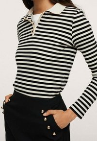 Polo a maniche lunghe a righe bianche e nere con collare beige, caratterizzato da un colletto con zip e dettagli in oro sui taschini.