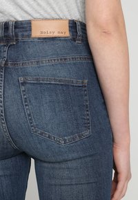 Jeans in denim blu scuro con design a vita media, caratterizzati da una patch in pelle marrone con l'etichetta "Noisy may" e tasche posteriori standard.