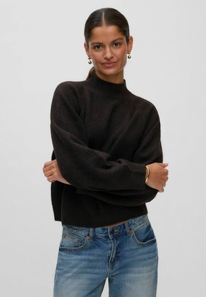 Kvinde med mørkt hår trukket tilbage bærer en sort sweater, blå jeans, guldringrøgøreringe og en guldbangle, stående med armene krydset.