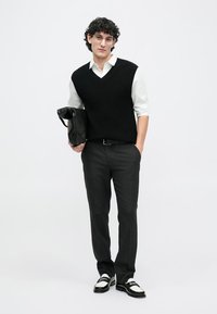 Young man with curly hair wearing glasses, white shirt, black sweater vest, black pants, carrying a black leather bag, standing.