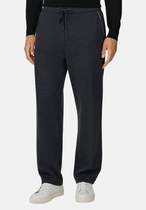 Mand iført mørkegrå snor-pants og hvide sneakers står med hænderne langs siden på lys baggrund.