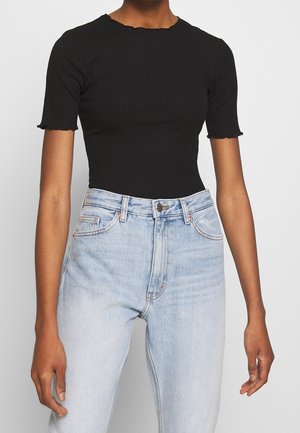 Person iført en tætsiddende sort ribbet kortærmet top og lyseblå high-waist denimjeans, stående mod en ensfarvet baggrund.