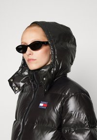 Chaqueta inflable negra con textura brillante, capucha y botones a presión. Presenta un colorido logo de Tommy Jeans en el pecho y elegantes gafas de sol negras.
