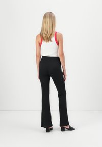 Femme blonde portant un haut blanc sans manches avec des bretelles rouges, un pantalon noir ajusté et des sandales noires à talons, debout de dos sur fond blanc.