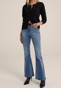 Cardigan nero a coste waffle, jeans a zampa d'elefante blu chiaro e stivali neri con tacco alto. Il cardigan presenta bottoni sul davanti, i jeans hanno un classico design a cinque tasche.