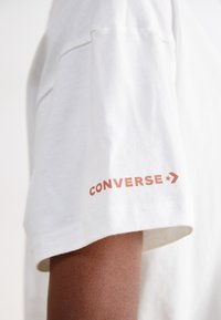 Valge puuvillane t-särk lühikeste varrukatega, millel on vasakul varrukal rustikavärvi "Converse" logo ja tähtede aktsent.