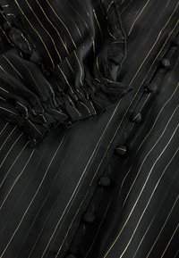 Tessuto nero con sottili righe verticali dorate, polsini elastici arricciati e una fila di bottoni coperti lungo la parte anteriore.