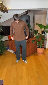 Brown long-sleeve shirt with a half-zip collar, paired with dark gray pants and metallic gray shoes, standing indoors near a plant.