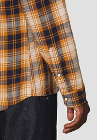 Mano di un uomo accanto a una camicia a maniche lunghe a quadri gialla, blu navy e bianca con bottoni a pressione, abbinata a pantaloni in denim scuri.