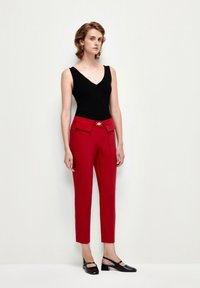 Pantalones tailored rojos con bolsillos laterales, con una hebilla de cinturón dorada. Combinados con una blusa negra sin mangas. Tejido suave, corte ajustado.