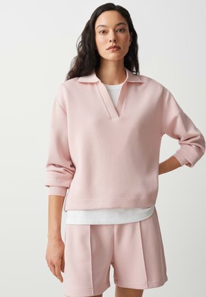 Frau trägt hellrosa langärmeliges Oberteil mit Kragen über weißem Hemd und passenden rosa Shorts, steht vor einem schlichten hellen Hintergrund.