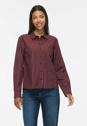 Femme portant une chemise bordeaux à manches longues avec des rayures verticales blanches et un jean bleu, souriant directement à la caméra.