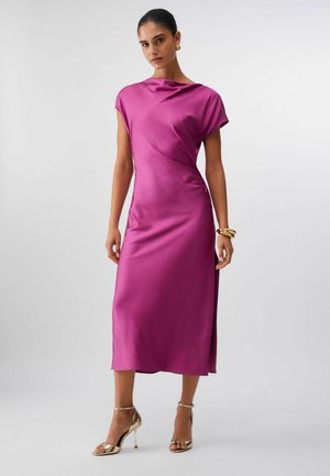 Ženska, oblečena v brezrokavno midi obleko iz magenta krhkega satenastega materiala, z zlatimi obročastimi uhani, zapestnicami in zlatimi sandali z visokimi petami, stoji pred enostavnim ozadjem.