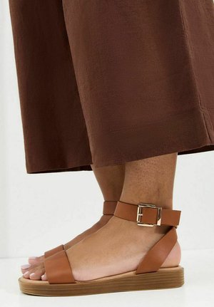 Voeten met bruine sandalen met enkelbandje, platte houten zolen en een brede voorkant, gecombineerd met bruine cropped broeken.