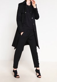 Černý trenčkot s knoflíky, vrstvený přes černý top a odpovídající kalhoty. Charakteristické jsou sandály s otevřenou špičkou a strukturovaný design.