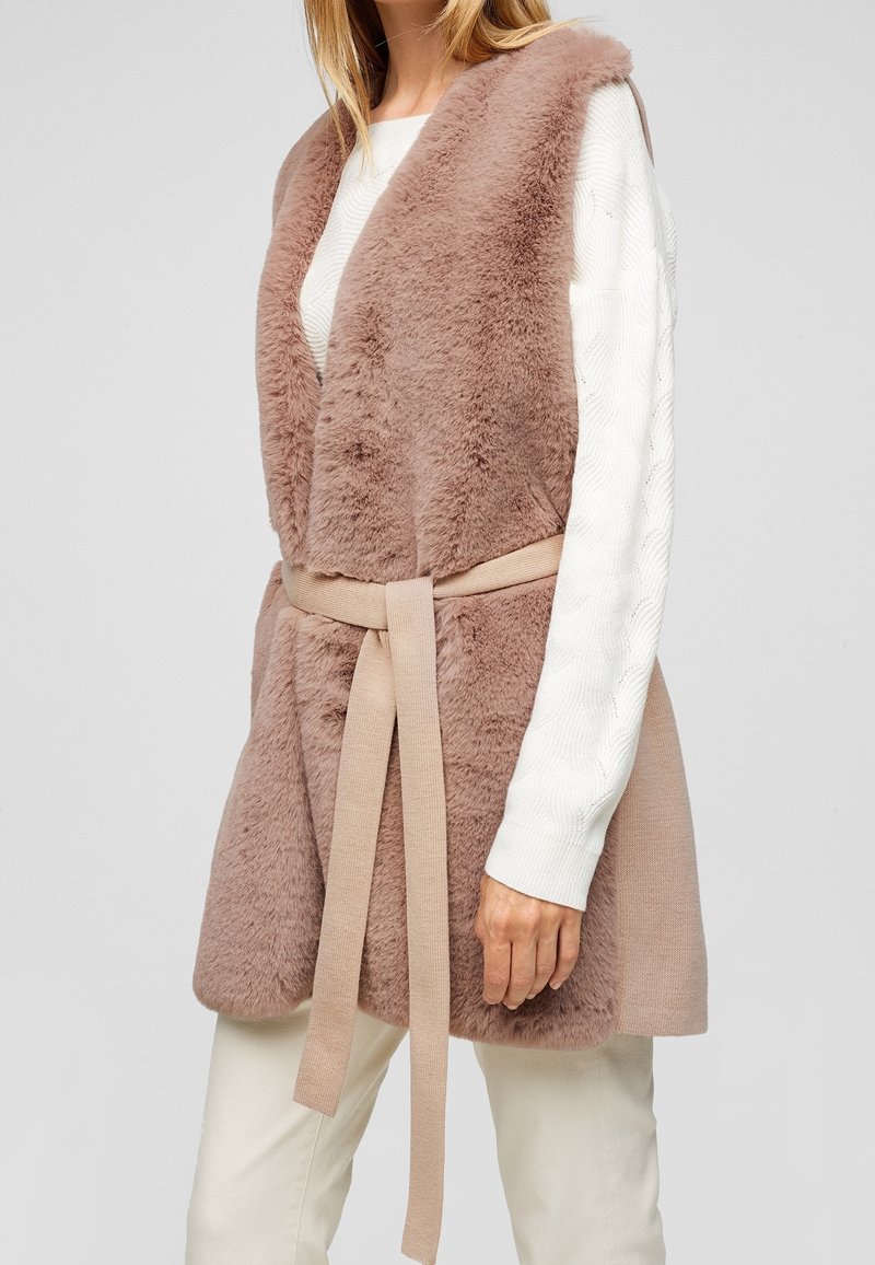 Femme portant un gilet sans manches en fausse fourrure beige avec une ceinture en tissu assortie, par-dessus un pull blanc à manches longues et un pantalon clair.