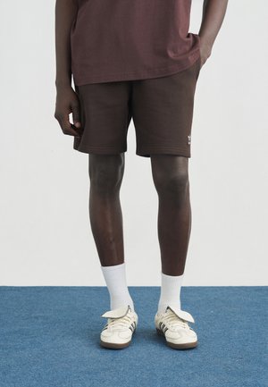Hombre con pantalones cortos marrones, calcetines blancos y zapatillas blancas de pie sobre una alfombra azul con las manos relajadas a los lados.