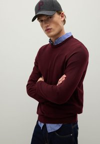 Maglione a maglia bordeaux sopra una camicia a righe blu e bianche, indossato con un cappellino grigio scuro con il logo rosso "P", con le braccia incrociate.