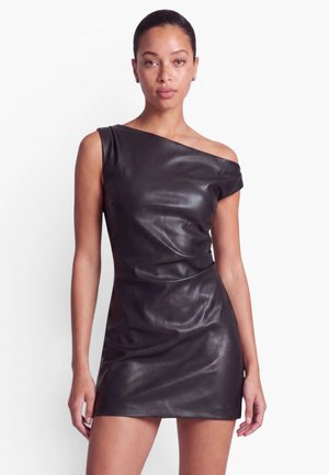 Femme portant une mini-robe en cuir noire ajustée avec une épaule dénudée, debout devant un fond blanc uni.