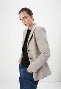 Světlé šedé sako na míru se dvěma předními knoflíky, nošeno přes černý top. Model má na sobě modré džíny, ruce v kapsách, na bílém pozadí.