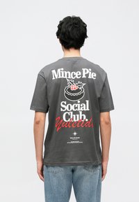 Sivé bavlnené tričko s krátkymi rukávmi s grafickým nápisom: „Mince Pie Social Club Yuletide“ a dekoratívnou ilustráciou koláča.