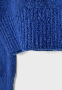 Gros plan d’un tissu tricoté bleu montrant une texture côtelée et des fibres douces avec un détail de couture pliée sur un fond uni.