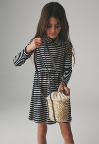 Flicka i en marinblå och vit randig klänning som håller i och ser ner i en liten vävd korg med en tygpås som har snöre i. 