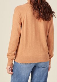 Světlě hnědý svetr s jemnou texturou, s půlkulatem a žebrovanými manžetami, kombinovaný s modrými džínami širokých nohavic.