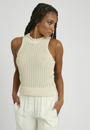 Débardeur en tricot beige avec un col montant, texture côtelée et design ajusté. Porté avec un pantalon large de couleur crème.