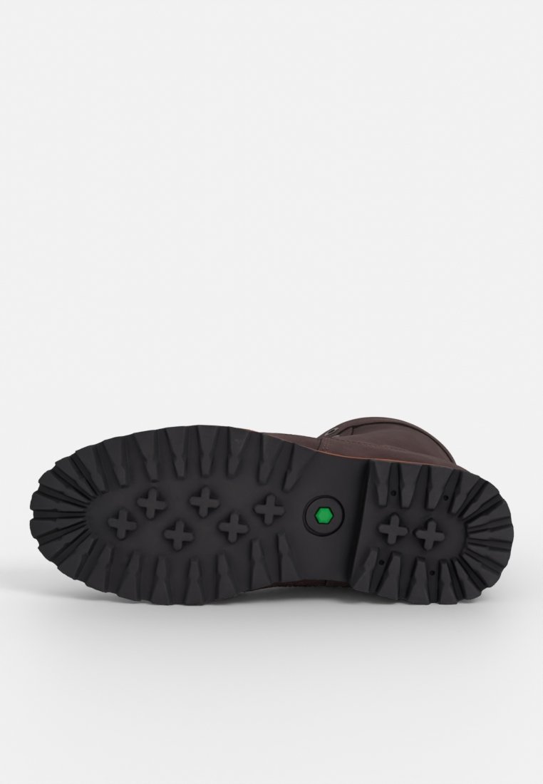 Suela de bota marrón con patrones de tracción profundos, material de goma y un detalle circular verde. Presenta un borde redondeado y una superficie de agarre texturizada.