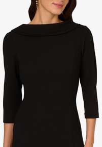 Kobieta w dopasowanej czarnej sukience z dekoltem w łódkę i rękawami do trzech czwartych, uzupełniona długimi kolczykami.