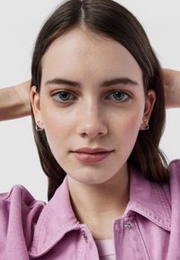 Orecchini d'argento con forma a contorno floreale. Il modello indossa una giacca di jeans rosa con cuciture visibili e bottoni a pressione. Texture della pelle liscia.
