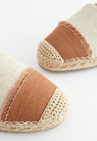 Vaaleanruskeat ja kermaiset espadrillot, joissa on pyöristetty kärki, teksturoitu kangasosa, kontrastivärinen varvaskärki ja punottu juuttireunus pohjan ympärillä.
