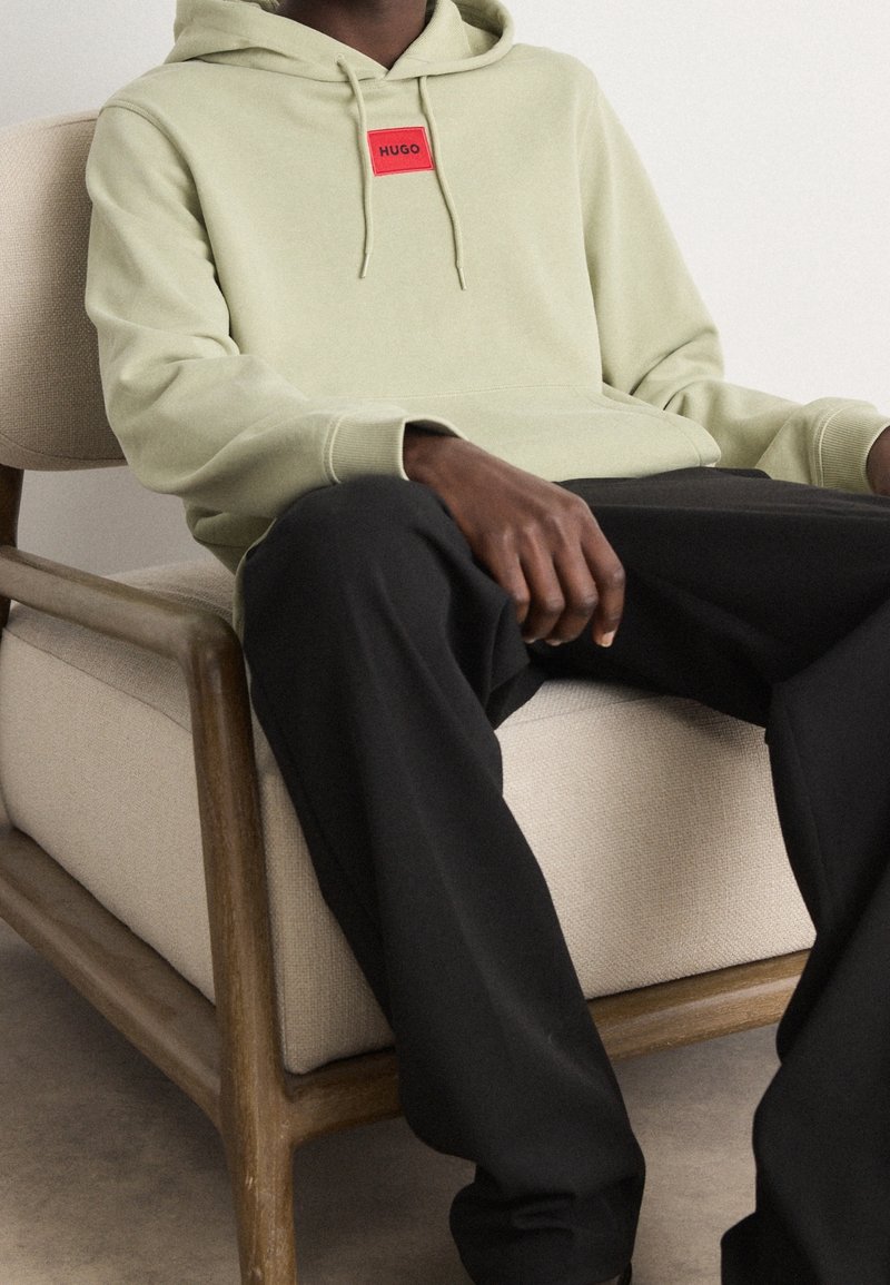 Personne assise portant un sweat à capuche beige avec un logo rouge "HUGO" et un pantalon noir sur une chaise en bois beige rembourrée.